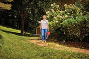 Person holding CRAFTSMAN cordless leaf blower CMCBL700D1 on garden pathway, surrounded by green foliage and flowers.