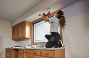 A person wearing protective glasses and a mask uses a CRAFTSMAN cordless drill to install or remove a fixture in the ceiling above a kitchen counter. The person is kneeling on the countertop, surrounded by cabinets and renovation materials.
