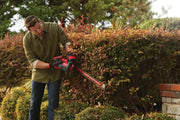 Photo of a person trimming hedges with a red CRAFTSMAN hedge trimmer, SKU CMCHTS820D1, outdoors in a garden.