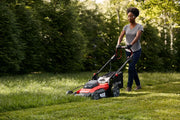Photo of a person using a Craftsman walk-behind lawn mower CMCMWSP220P2, cutting grass in a yard with trees in background.