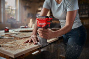 Photo of a person using a Craftsman cordless compact router, SKU CMCW400B, on a wooden board in a workshop.