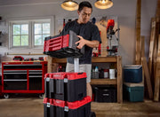 A person placing a red and black CRAFTSMAN tool box onto a stack of similar tool boxes in a garage workshop. The tool boxes are part of a modular storage system, and the workspace features various tools, a workbench, and storage cabinets.