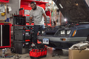 A person working on a car with its hood open in a garage. There are multiple toolboxes and a red tool bag filled with tools nearby, as well as various automotive parts and equipment on the floor and shelves.