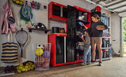 A garage scene featuring CRAFTSMAN red and black storage cabinets and shelving units used to organize various sports equipment, toys, and household items. Two people are standing near the cabinets, with the adult reaching for an item and the child looking inside. The area appears tidy and well-organized.
