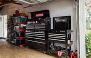 CRAFTSMAN branded black tool chests and rolling tool carts with multiple drawers are organized in a garage workspace. Shelving units with bins and various garage items are visible beside the tool chests.