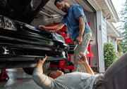 Two people working together on a car in a garage. One person is lying on the floor under the car, reaching up, while the other stands nearby holding a tool. The car's hood is open and there is a red tool chest in the background.