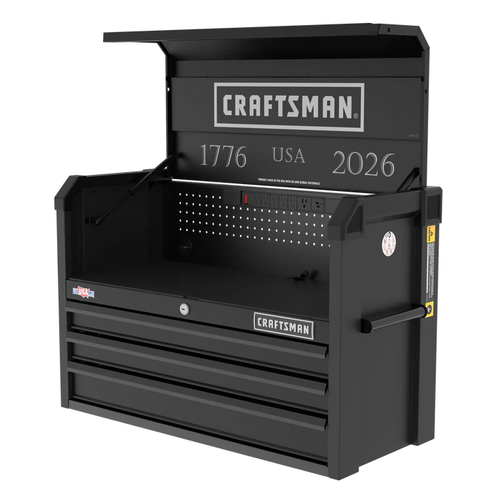 A black Craftsman-branded metal tool chest with three drawers, a hinged top lid, side handle, and built-in power strip. The interior back panel features the text '1776 USA 2026' and a perforated panel for organizing tools.