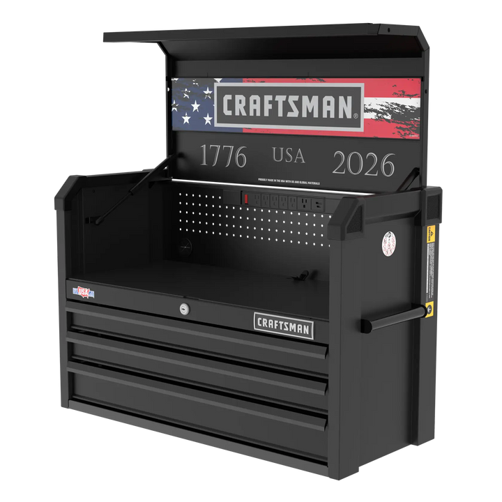 A black Craftsman tool chest with three drawers, an open top compartment, and a built-in pegboard. The chest features an American flag design, the years '1776' and '2026', and 'USA' displayed inside the lid. The tool chest also includes a set of electrical outlets built into the back panel.