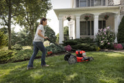 Photo of a person using a red Craftsman walk-behind lawn mower CMXGMAM2703840 on a grassy yard in front of a house.
