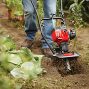Photo of Craftsman tiller CMXGVAMKC25C turning soil in a garden with leafy vegetables.