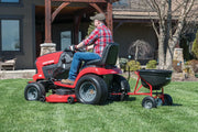 Photo of Craftsman lawn tractor towing black spreader, SKU CMXGZBF7124571, on grass lawn, side angle.