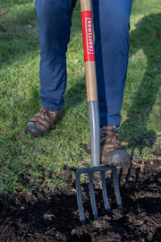 Photo of a Craftsman CMXMLTP35327 garden fork with wood handle in use on soil, shown outdoors with person wearing boots.