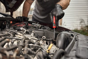 A person wearing black gloves uses a red-handled CRAFTSMAN screwdriver to work on the engine of a car with the hood open.