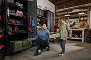 A woman kneeling next to an open storage cabinet with organized tools and equipment, while a child stands beside her holding
